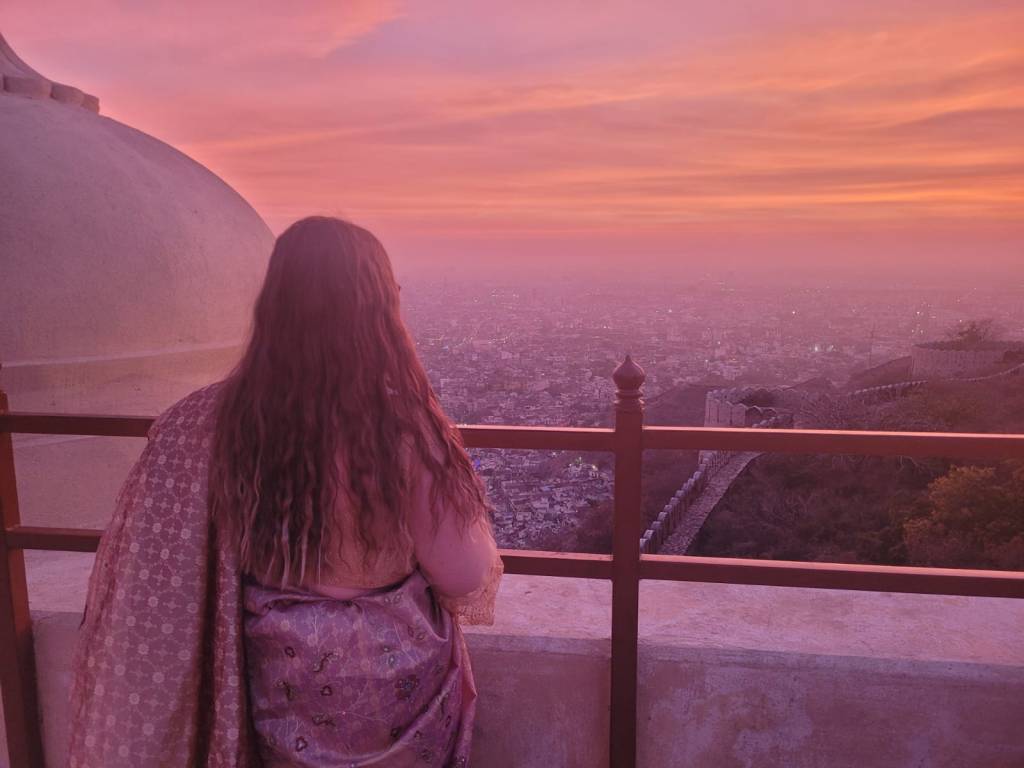 Image of woman wearing a saree with her back to the camera with pink and orange sunset in the background over the city